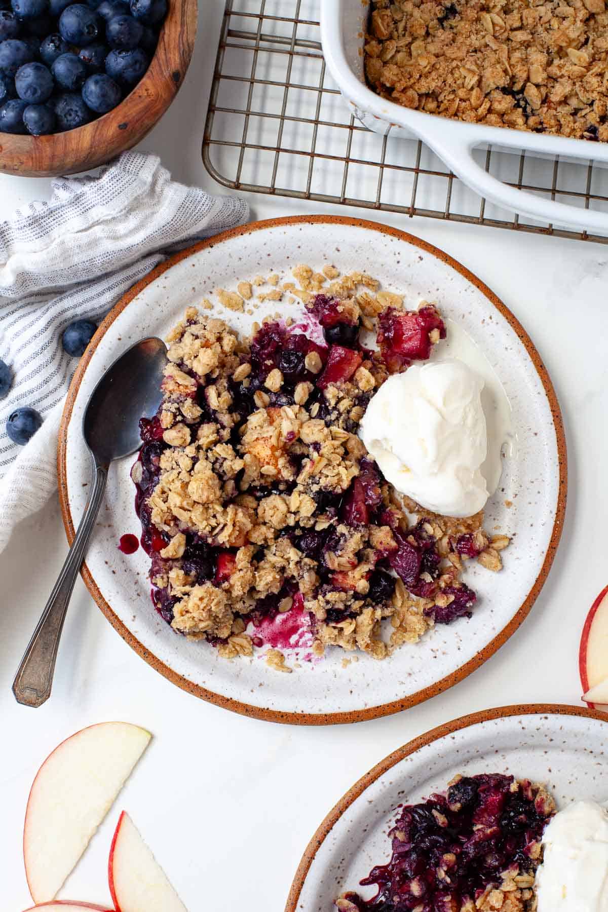 Two plates of blueberry apple crisp served with vanilla ice cream next to baking dish with crisp